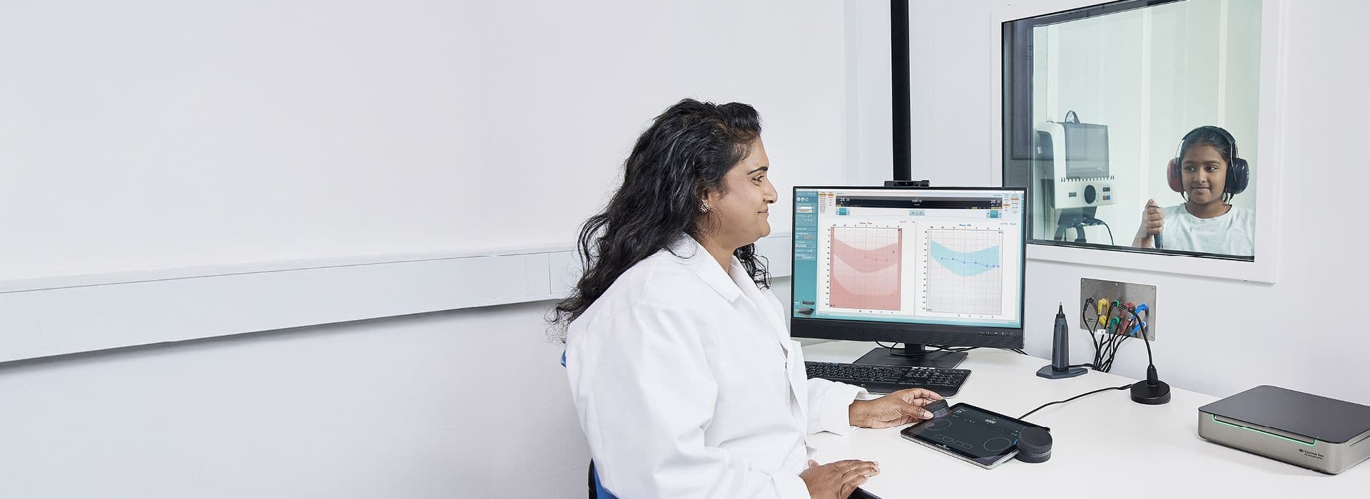 En pige sidder i en lydisoleret kabine og gennemgår en høretest udført af en audiolog, der betjener Touch Keyboardet på Equinox Evo.